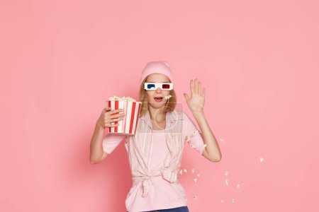 Foto de Aterrorizada chica con corte de pelo corto viendo película de terror, sosteniendo cubo de palomitas de maíz sobre fondo rosa - Imagen libre de derechos
