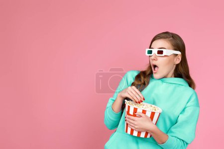 Foto de Asustada mujer morena rizada aterrorizada en sudadera viendo película, sosteniendo cubo de palomitas de maíz sobre fondo rosa - Imagen libre de derechos