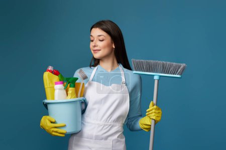 Foto de Mujer sonriente con guantes de goma amarillos y delantal más limpio sosteniendo cubo de detergentes y escoba sobre fondo azul. - Imagen libre de derechos