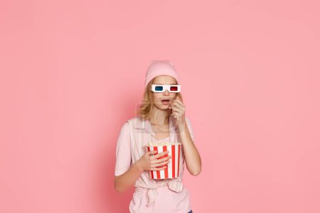 Foto de Aterrorizada chica con corte de pelo corto viendo película de terror, sosteniendo cubo de palomitas de maíz sobre fondo rosa - Imagen libre de derechos