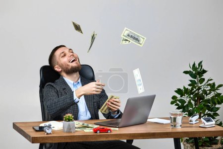 Téléchargez les photos : Young bearded businessman working on laptop and throwing money while sitting on chair at desk. banknotes fly in the air - en image libre de droit