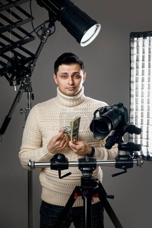 Photo for Professional handsome photographer with digital camera on tripod holding hundred dollar bills on gray background with lighting equipment - Royalty Free Image