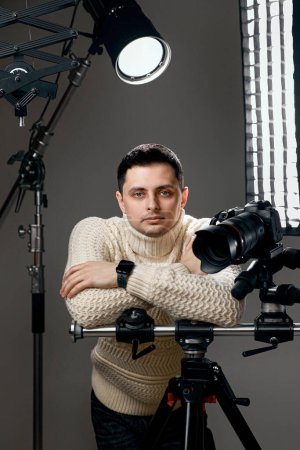 Téléchargez les photos : Photographe professionnel avec appareil photo numérique sur trépied regardant l'appareil photo sur fond gris avec équipement d'éclairage - en image libre de droit