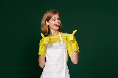Téléchargez les photos : Belle femme blonde en gants de caoutchouc jaune et tablier plus propre montrant ok signe sur fond vert. - en image libre de droit