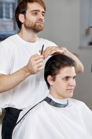 Téléchargez les photos : Coiffeur professionnel homme peignage homme caucasien assis sur la chaise avant la coupe de cheveux au salon de coiffure. client dit quelle coupe de cheveux il veut - en image libre de droit