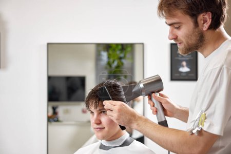 Téléchargez les photos : Coiffeur professionnel utilisant sèche-cheveux et peigne dans le salon de coiffure. Coupe de cheveux dans le salon de coiffure. - en image libre de droit