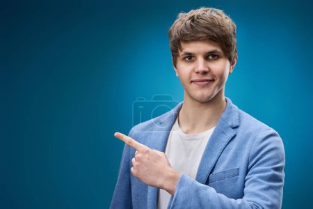Foto de Positivo caucásico joven hombre en chaqueta señalando el dedo en el espacio de copia sobre fondo azul - Imagen libre de derechos