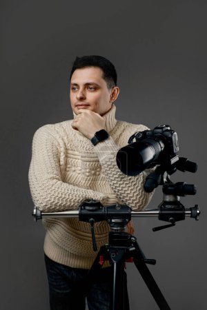Foto de Profesional joven fotógrafo guapo en suéter cerca de la cámara digital en trípode sobre fondo gris - Imagen libre de derechos