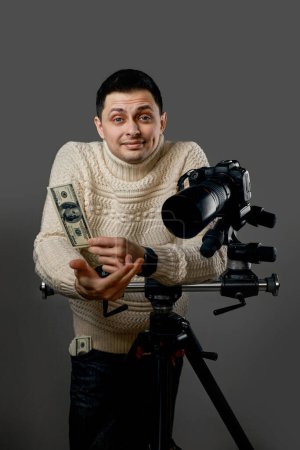 Téléchargez les photos : Photographe professionnel avec appareil photo numérique sur trépied tenant des billets de cent dollars sur fond gris - en image libre de droit