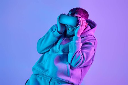Foto de Man in sweatshirt using virtual reality glasses and playing video games on blue background. Neon lighting - Imagen libre de derechos