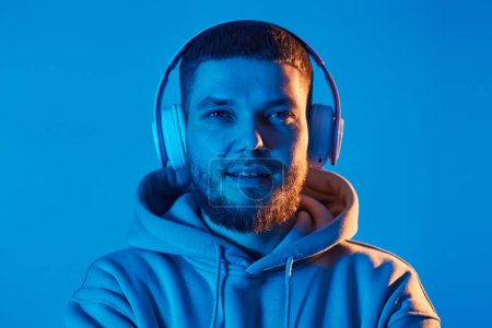 Foto de Handsome bearded man with headphones in sweatshirt enjoying favorite music on blue neon background. Neon lighting - Imagen libre de derechos