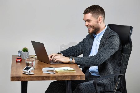 Foto de Hombre de negocios barbudo feliz escribiendo en el teclado de la computadora, enviando correos electrónicos a los socios de negocios, sentado en la silla en el escritorio - Imagen libre de derechos