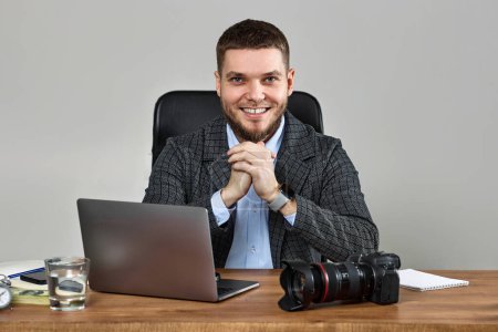 Photo for Successful bearded male photographer smiling cheerfully while working at desk. dslr camera on table - Royalty Free Image