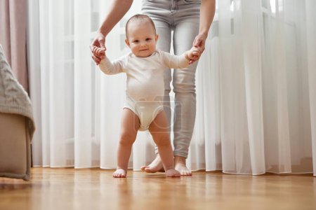 Photo for Happy little baby girl learning to walk with mother help at home. child take her first steps - Royalty Free Image