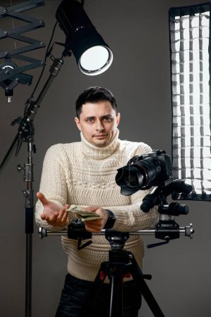 Foto de Fotógrafo caucásico sorprendido con cámara digital en trípode sosteniendo efectivo sobre fondo gris con equipo de iluminación - Imagen libre de derechos