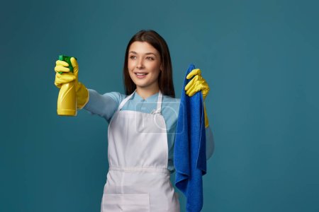 Foto de Mujer joven feliz en guantes de goma y delantal más limpio sosteniendo trapo de limpieza y rociador de detergente sobre fondo azul. centrarse en las manos - Imagen libre de derechos