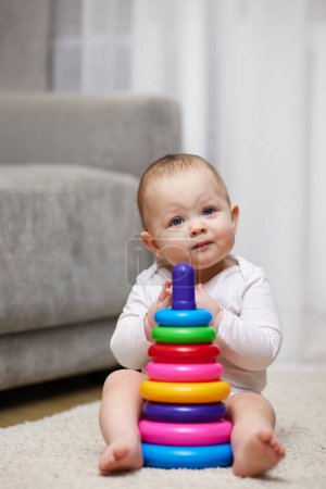 Téléchargez les photos : Bébé fille mignonne jouant avec la pyramide colorée jouet assis sur le tapis à la maison. Développement précoce pour les enfants. - en image libre de droit