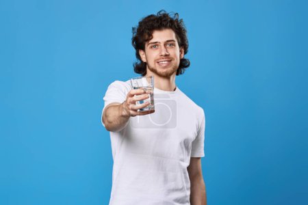 Téléchargez les photos : Heureux bel homme dans un t-shirt blanc montrant verre d'eau potable sur fond bleu. focus sur le verre - en image libre de droit