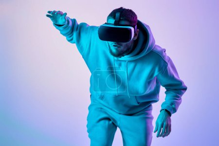 Téléchargez les photos : Homme en sweat-shirt en utilisant des lunettes de réalité virtuelle et de jouer à des jeux vidéo sur fond bleu. Éclairage au néon. jeu et divertissement en ligne - en image libre de droit