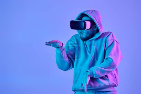 Téléchargez les photos : Man in sweatshirt using virtual reality glasses and playing video games on blue background. Neon lighting - en image libre de droit