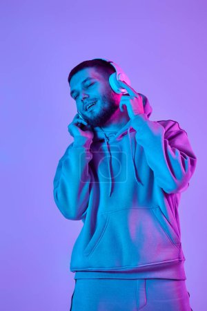 Téléchargez les photos : Handsome bearded man with headphones in sweatshirt enjoying favorite music on purple neon background. Neon lighting - en image libre de droit