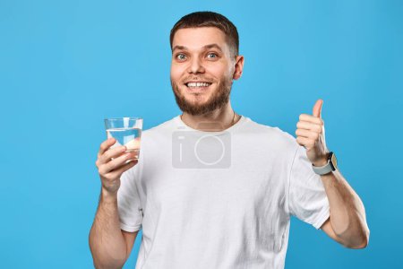 Foto de Hombre barbudo guapo en camiseta blanca con vaso de agua mostrando los pulgares hacia arriba gesto sobre fondo azul - Imagen libre de derechos
