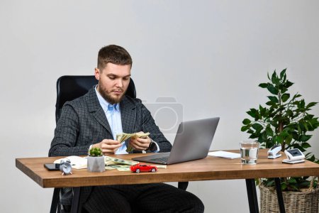 Photo for Young bearded businessman works on laptop and counts money at office desk - Royalty Free Image
