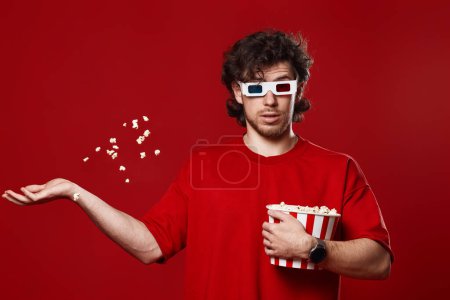 Foto de Joven sorprendido hombre en gafas 3d, camiseta roja vomitando palomitas de maíz sobre fondo rojo - Imagen libre de derechos