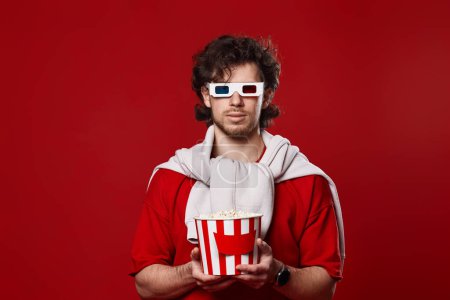 Photo for Young bearded man in 3d-glasses holding big bucket of popcorns on red background - Royalty Free Image