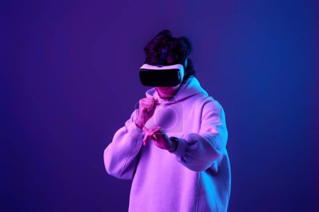 Foto de Man in sweatshirt using virtual reality glasses and playing video games on blue background. Neon lighting - Imagen libre de derechos