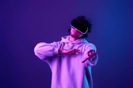 Foto de Man in sweatshirt using virtual reality headset on blue background. Neon lighting - Imagen libre de derechos