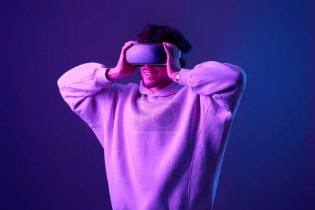 Photo for Man in sweatshirt using virtual reality headset on blue background. Neon lighting - Royalty Free Image