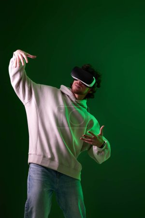 Téléchargez les photos : Man in vr glasses and sweatshirt playing video games with his friends on green background. Neon lighting - en image libre de droit