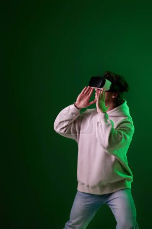 Téléchargez les photos : Man in sweatshirt using virtual reality headset on green background. Neon lighting - en image libre de droit