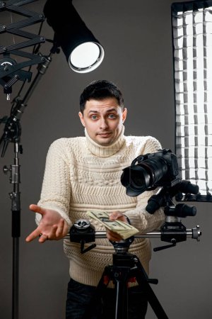 Téléchargez les photos : Photographe surpris avec appareil photo numérique sur trépied tenant de l'argent sur fond gris avec équipement d'éclairage - en image libre de droit