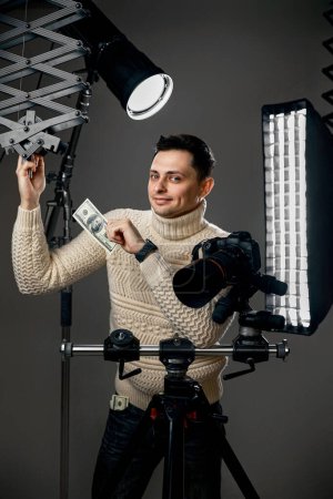 Téléchargez les photos : Professional handsome photographer with digital camera on tripod holding hundred dollar bills on gray background with lighting equipment - en image libre de droit