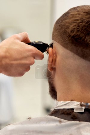 Foto de Peluquero recortar el cabello con clipper en el hombre barbudo guapo en la peluquería. - Imagen libre de derechos