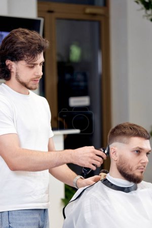 Photo for Barber trim hair with clipper on handsome bearded man in barber shop. - Royalty Free Image