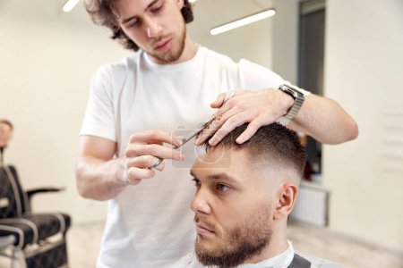 Foto de Hairdresser does haircut for caucasian bearded man using comb and grooming scissors in barber shop. - Imagen libre de derechos