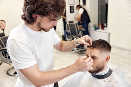 Téléchargez les photos : Hairdresser does haircut for caucasian bearded man using comb and grooming scissors in barber shop. - en image libre de droit