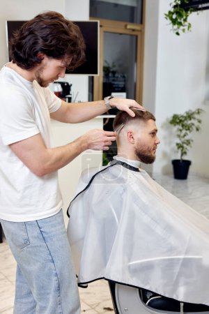 Photo for Barber trim hair with clipper on handsome bearded man in barber shop. - Royalty Free Image