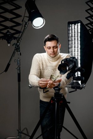 Foto de Fotógrafo guapo profesional con cámara digital en trípode sosteniendo efectivo sobre fondo gris con equipo de iluminación - Imagen libre de derechos