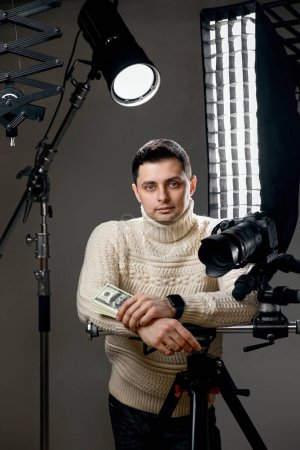 Photo for Professional handsome photographer with digital camera on tripod holding hundred dollar bills on gray background with lighting equipment - Royalty Free Image