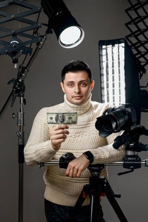 Téléchargez les photos : Beau photographe professionnel avec appareil photo numérique sur trépied montrant billet de cent dollars sur fond gris avec équipement d'éclairage - en image libre de droit