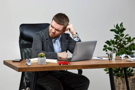 Foto de Bearded man sitting on chair at desk and suffering from headache at workplace, using laptop - Imagen libre de derechos
