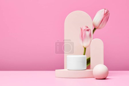 Téléchargez les photos : Pot blanc de produits cosmétiques haut de gamme sur podium avec tulipes roses et arc élégant sur fond rose pastel. maquette - en image libre de droit