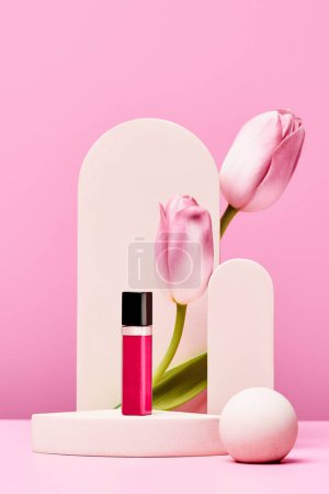 Foto de Brillo de labios en el podio con tulipanes rosados y elegante arco sobre fondo rosa pastel. diseño de moda minimalista. burla para arriba - Imagen libre de derechos