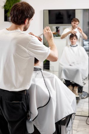 Téléchargez les photos : Young caucasian man getting haircut by professional male hairstylist using comb and grooming scissors at barber shop. - en image libre de droit