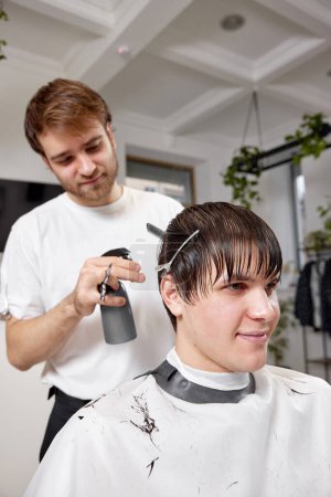 Téléchargez les photos : Homme heureux se faire couper les cheveux par professionnel coiffeur masculin en utilisant peigne et pulvérisateur au salon de coiffure. - en image libre de droit