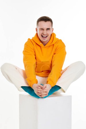 Foto de Hombre joven con sudadera naranja sentado en cubo blanco sobre fondo blanco. Longitud completa - Imagen libre de derechos